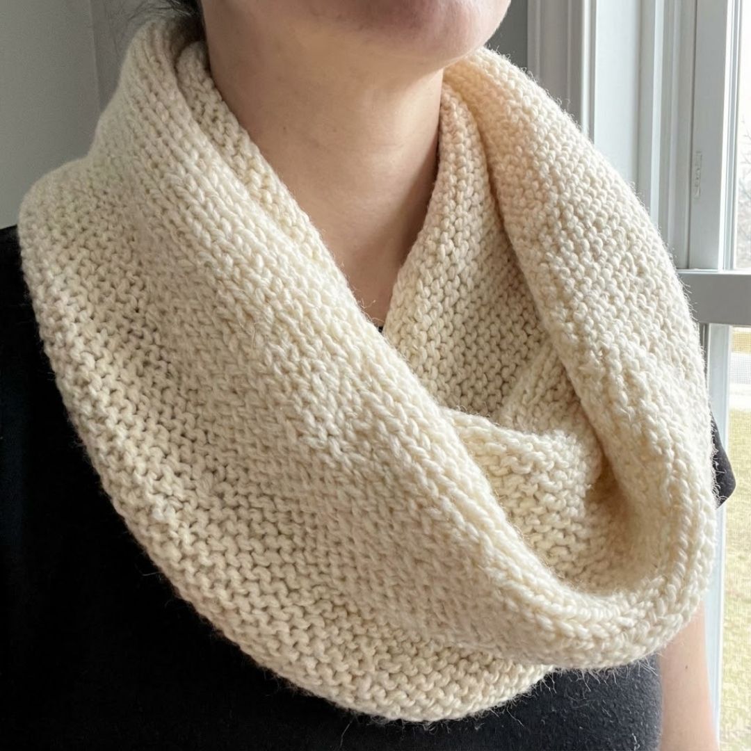 A person wearing a finished, cream-colored knit cowl with a simple garter stitch texture, draped casually