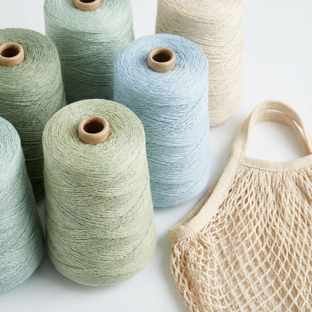 A collection of cotton and linen yarn on cones in light, fresh colors, next to a woven market bag